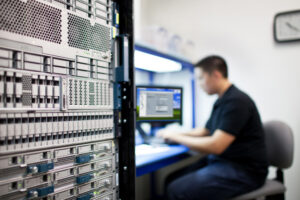 IT technician sits at a workstation beside a tall server rack, monitoring a monitor in a data center environment.