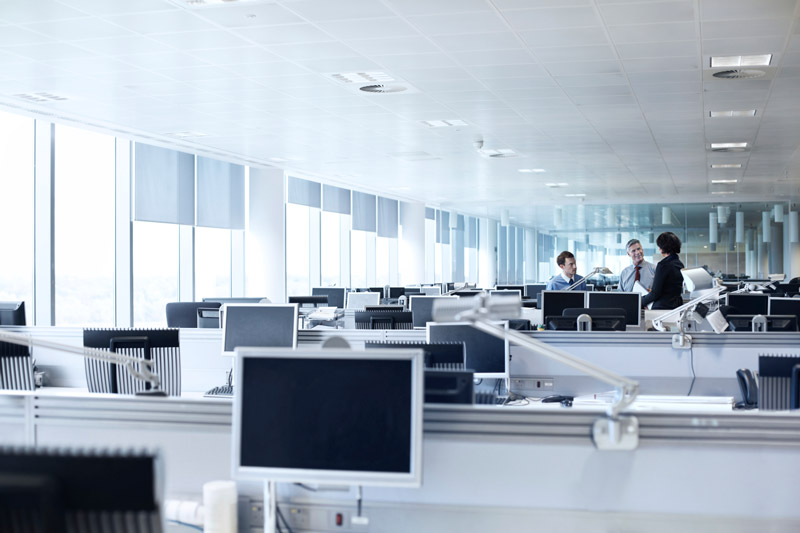 An open-plan office with rows of desks and computer monitors; a small group of three colleagues chatting at the far end.