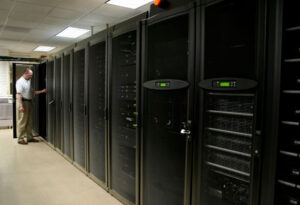 technician_working - Helpdesk Informática - Helpdesk Informática IT technician in a white shirt stands by a row of tall black server racks in a data center corridor, door open to a cabinet.